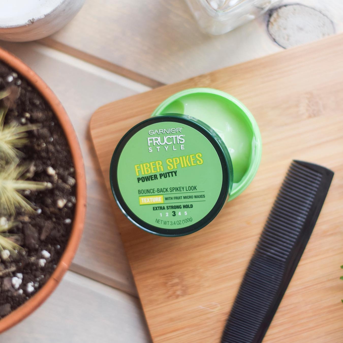 Garnier Fructis Style Fiber Spikes Power Putty opened on a wooden cutting board with a black comb on a distressed gray-painted wood table with a potted plant, a rectangular glass jar, and an empty white pottery piece.
